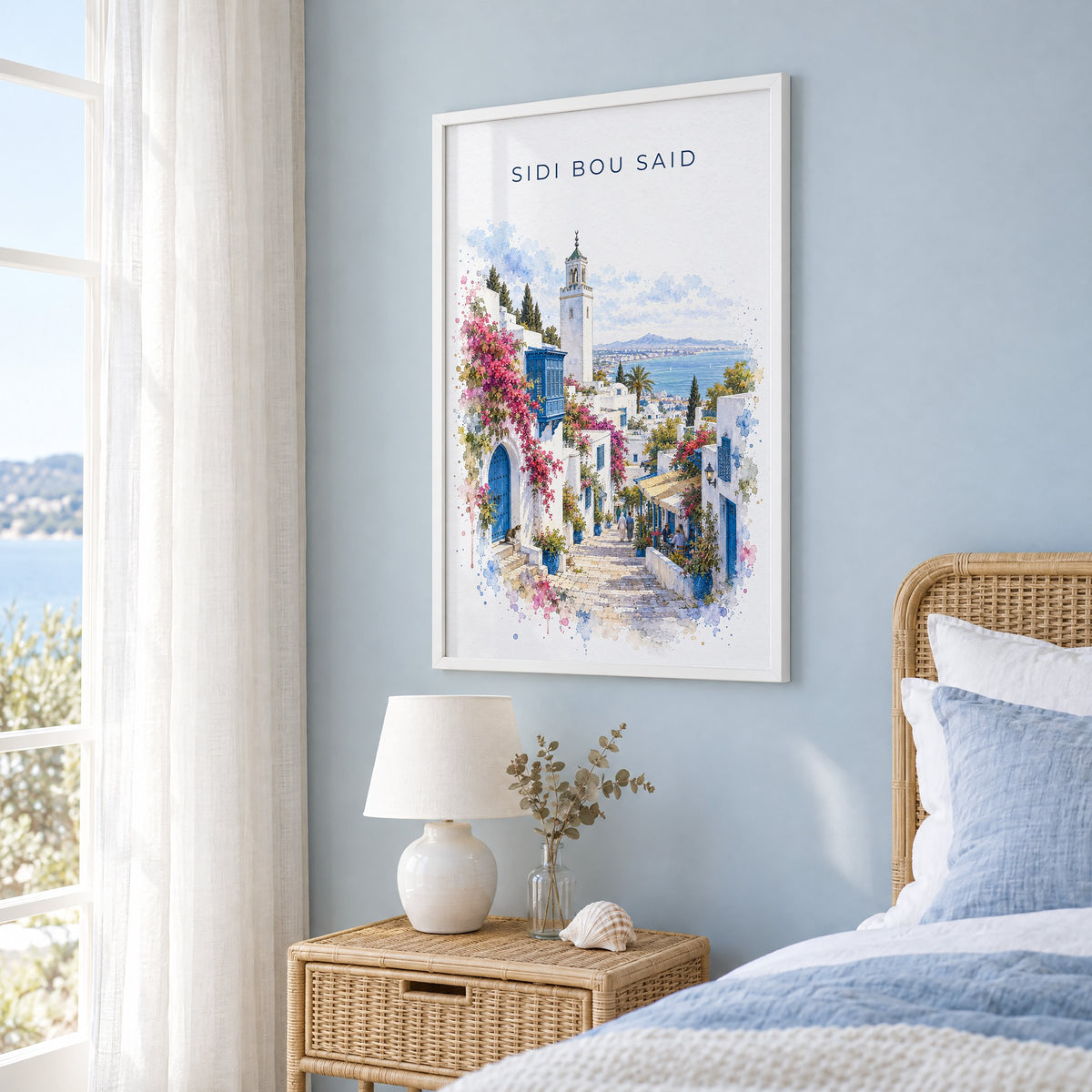 Sidi Bou Said watercolor print in a white frame on a pale blue wall beside a rattan bed in a coastal bedroom