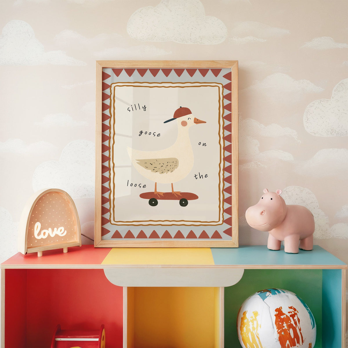 Silly goose on the loose nursery print in an oak frame on a rainbow toy shelf against cloud wallpaper
