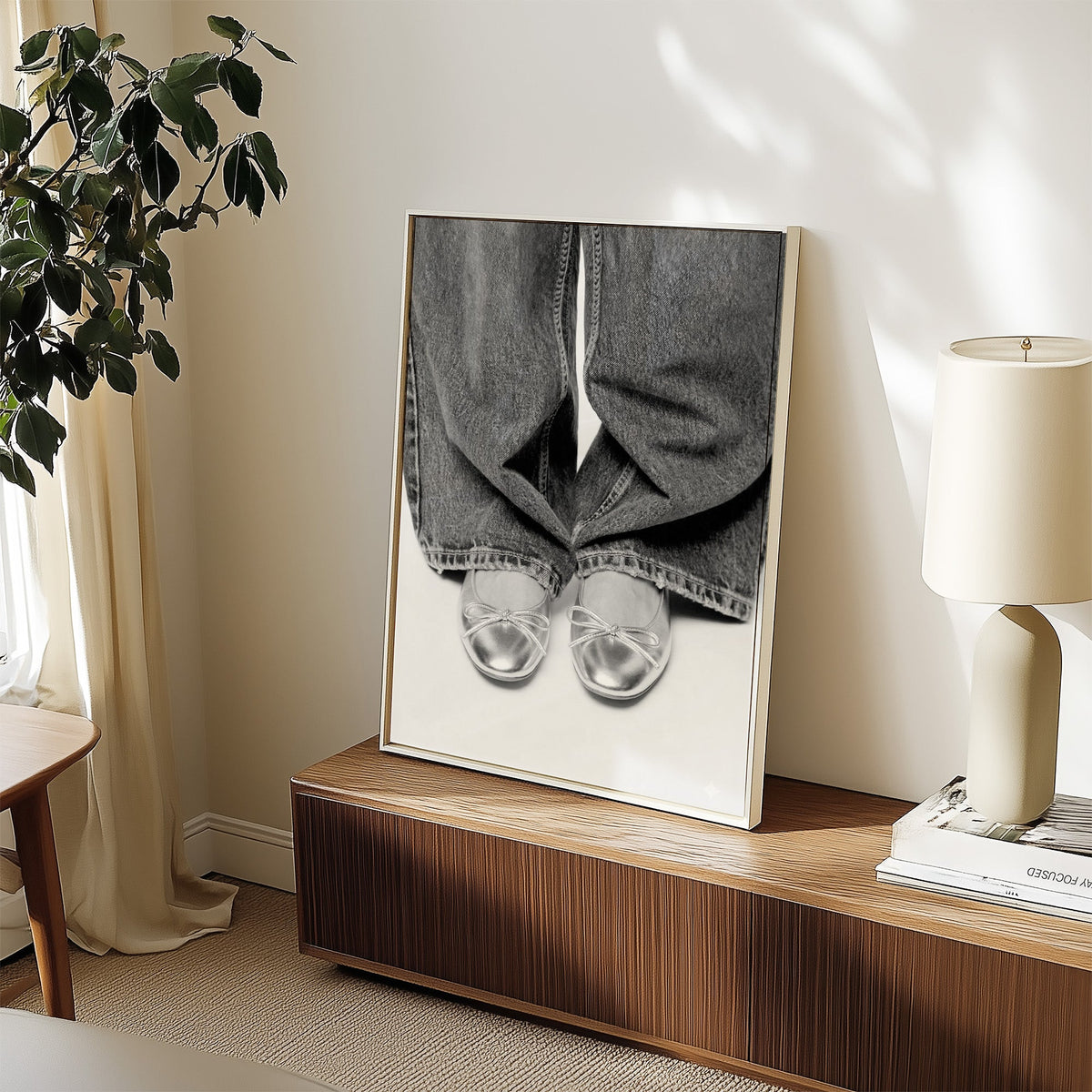 Silver ballet flats under wide-leg jeans print in a white frame on a walnut sideboard beside a ceramic lamp