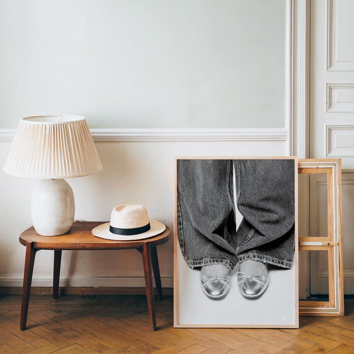 Silver ballet flats under wide-leg jeans print in a pine frame on a wood floor beside a lamp and straw hat