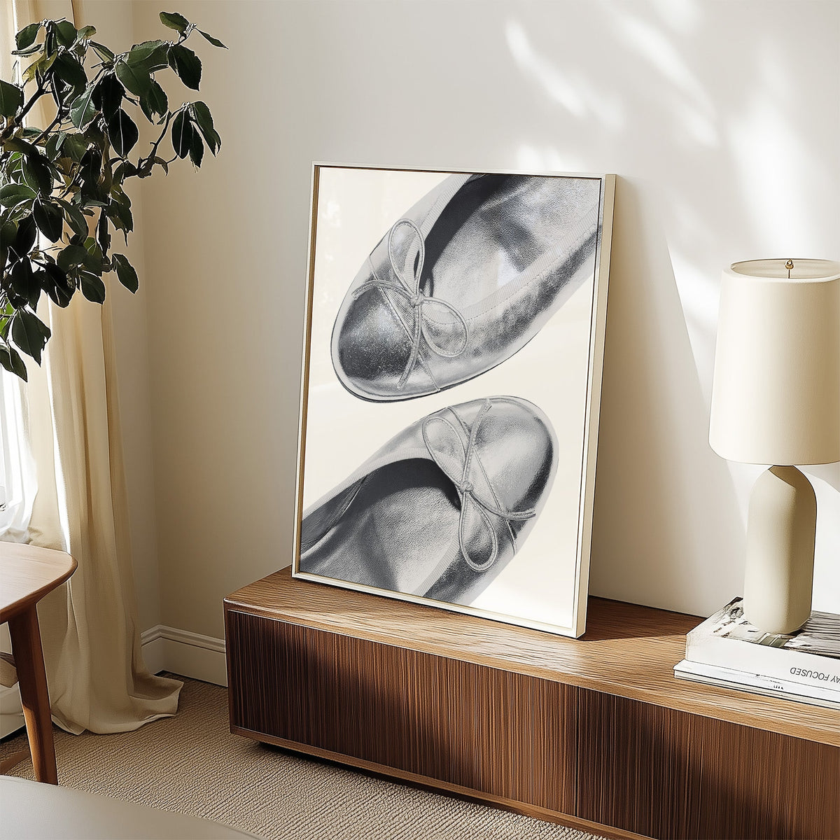 Plain toe silver ballet flats from above print in a white frame on a walnut sideboard beside a ceramic lamp