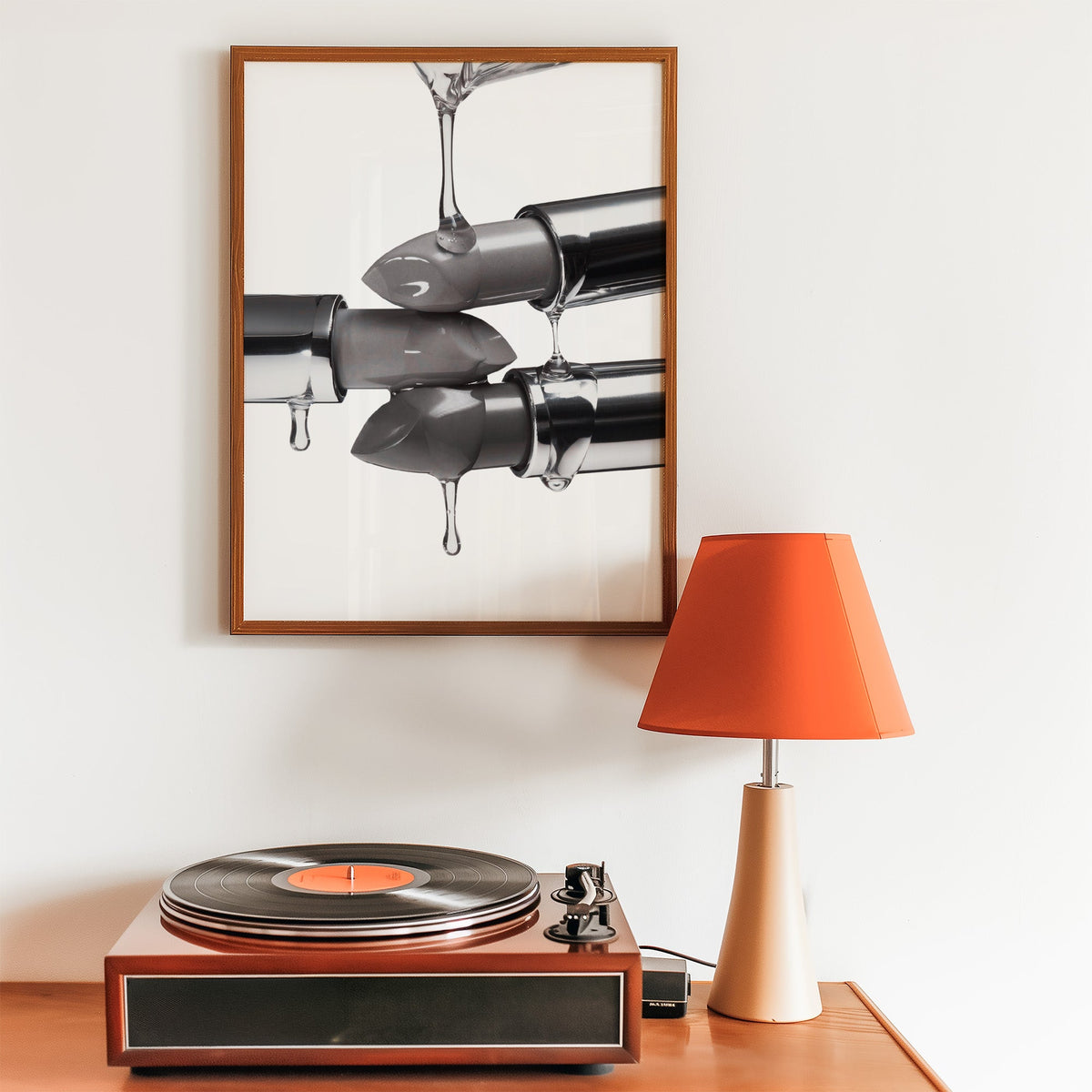 Silver lipsticks with clear gel drips print in a walnut frame above a record player and orange lamp