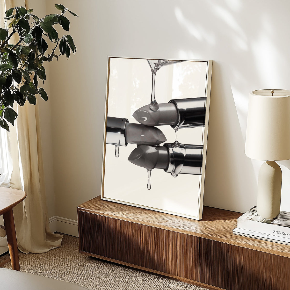 Silver lipsticks with clear gel drips print in a white frame on a walnut sideboard beside a ceramic lamp