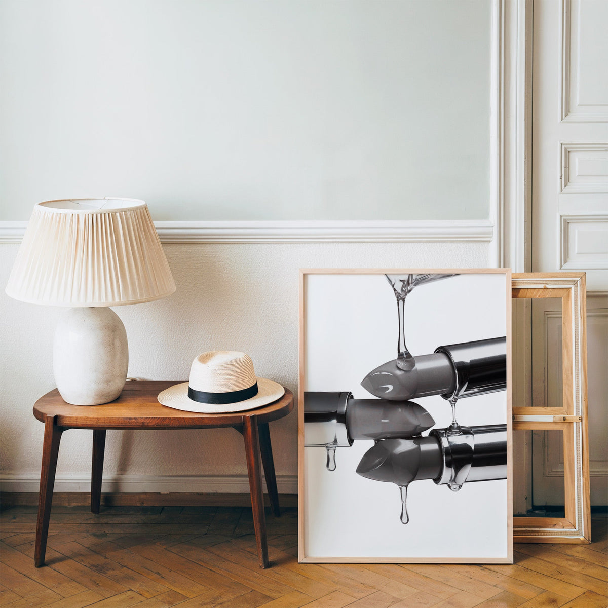 Silver lipsticks with clear gel drips print in a pine frame on a wood floor beside a lamp and straw hat