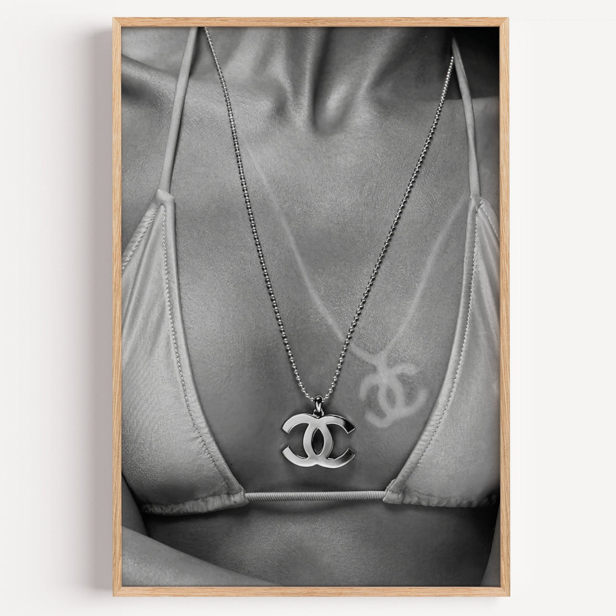 Close-up of logo pendant necklace and bikini tan line print in a light oak frame on a plain white wall