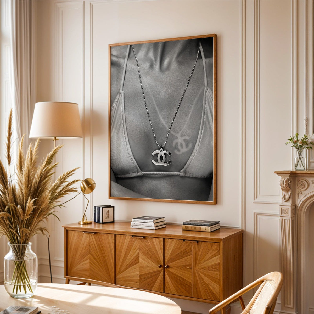 Close-up of logo pendant necklace and bikini tan line print in an oak frame above a sunburst wood sideboard with pampas grass