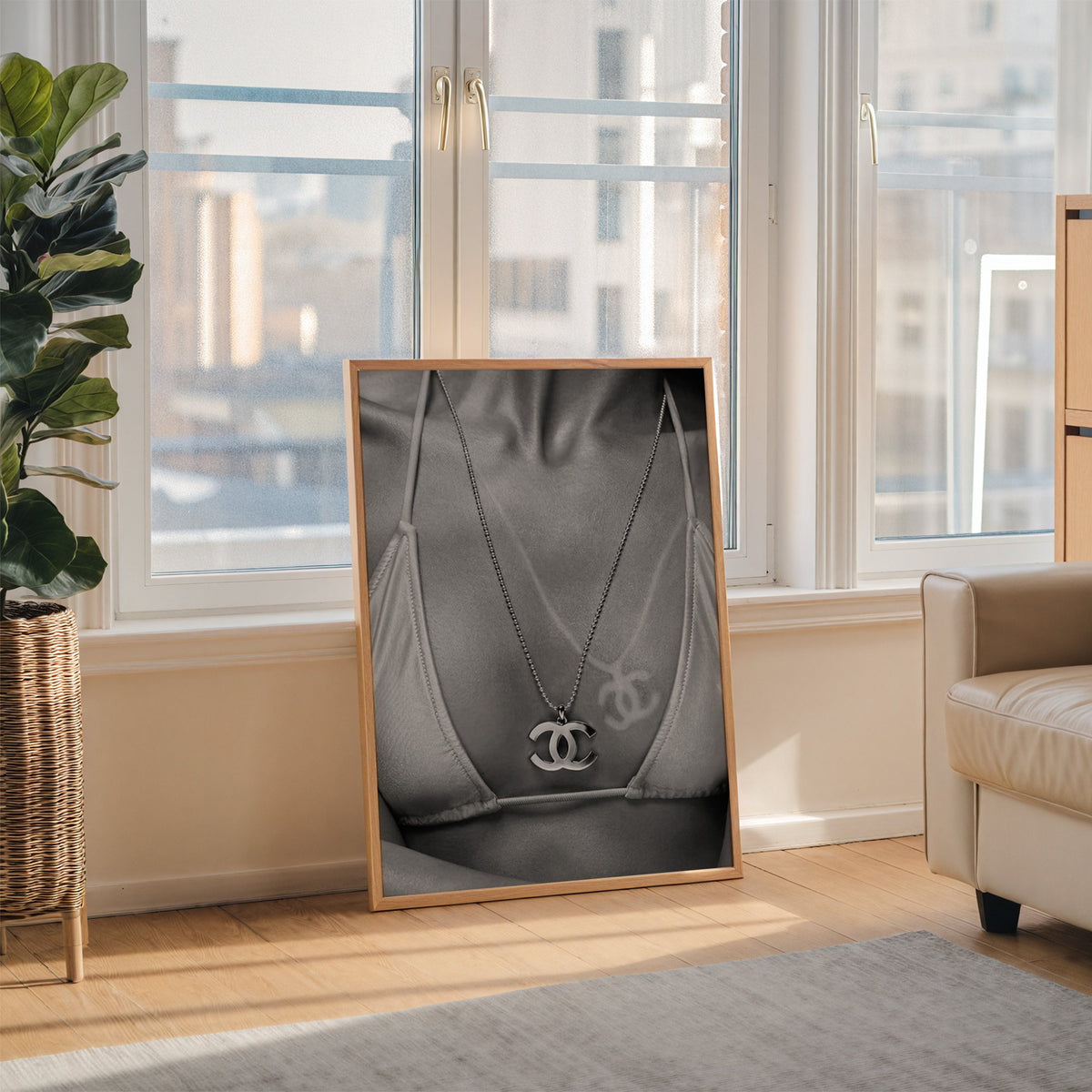 Close-up of logo pendant necklace and bikini tan line print in an oak frame leaning on the floor beside a fiddle leaf fig