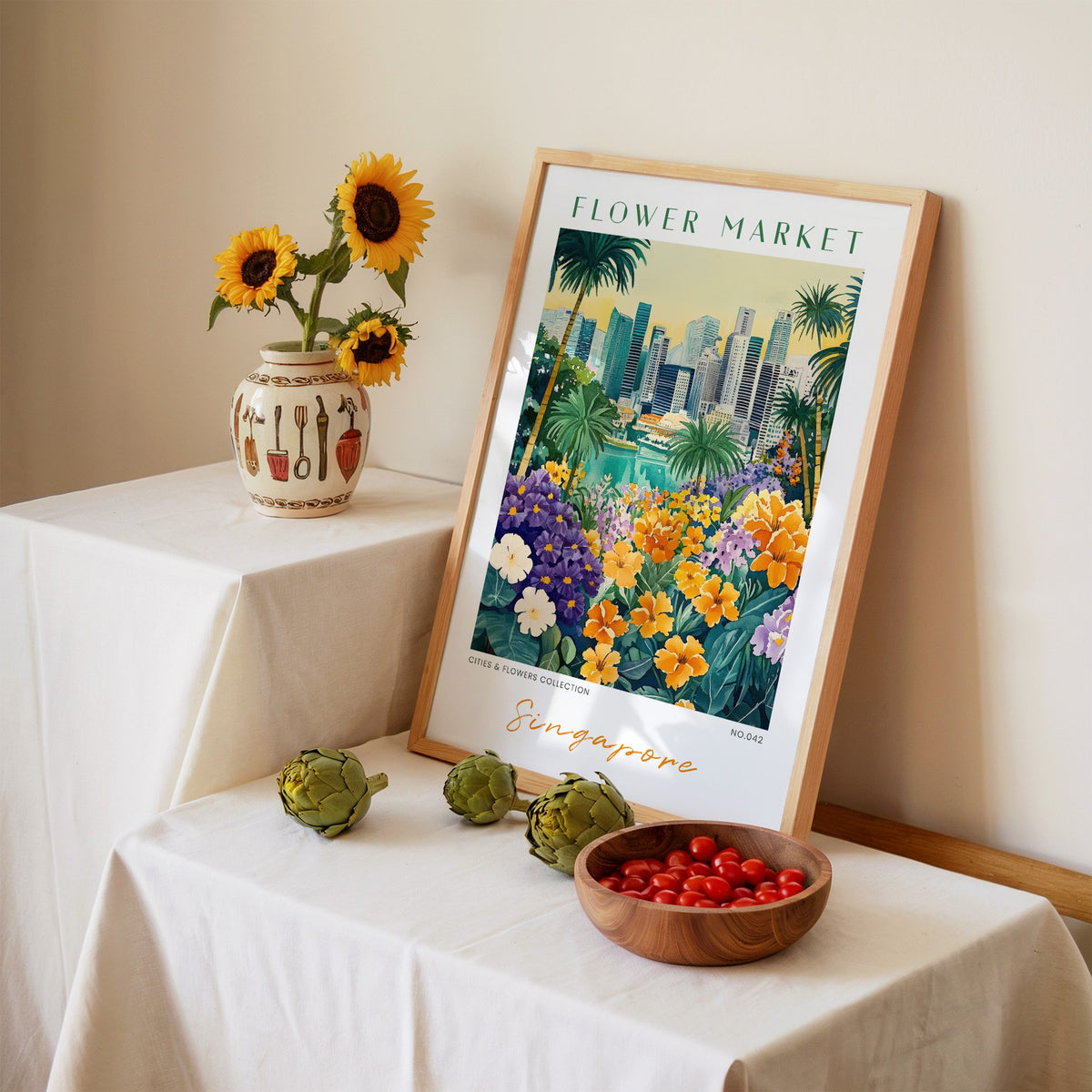Singapore flower market print in an oak frame leaning on a linen-draped table beside a vase of sunflowers