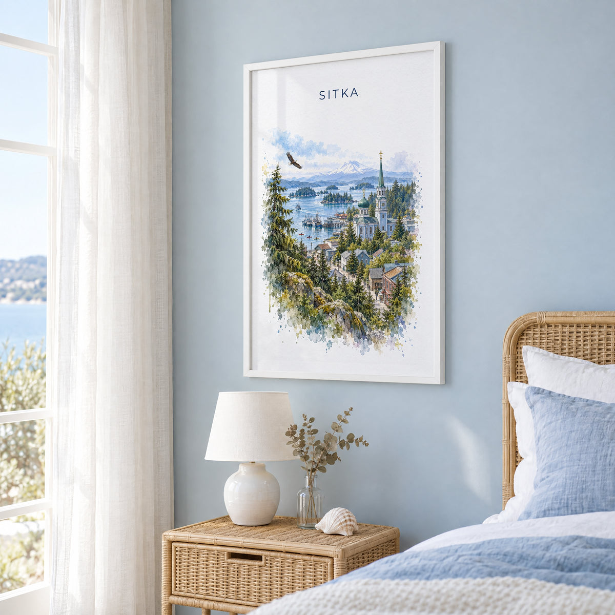 Sitka, Alaska watercolor print in a white frame on a pale blue wall beside a rattan bed in a coastal bedroom
