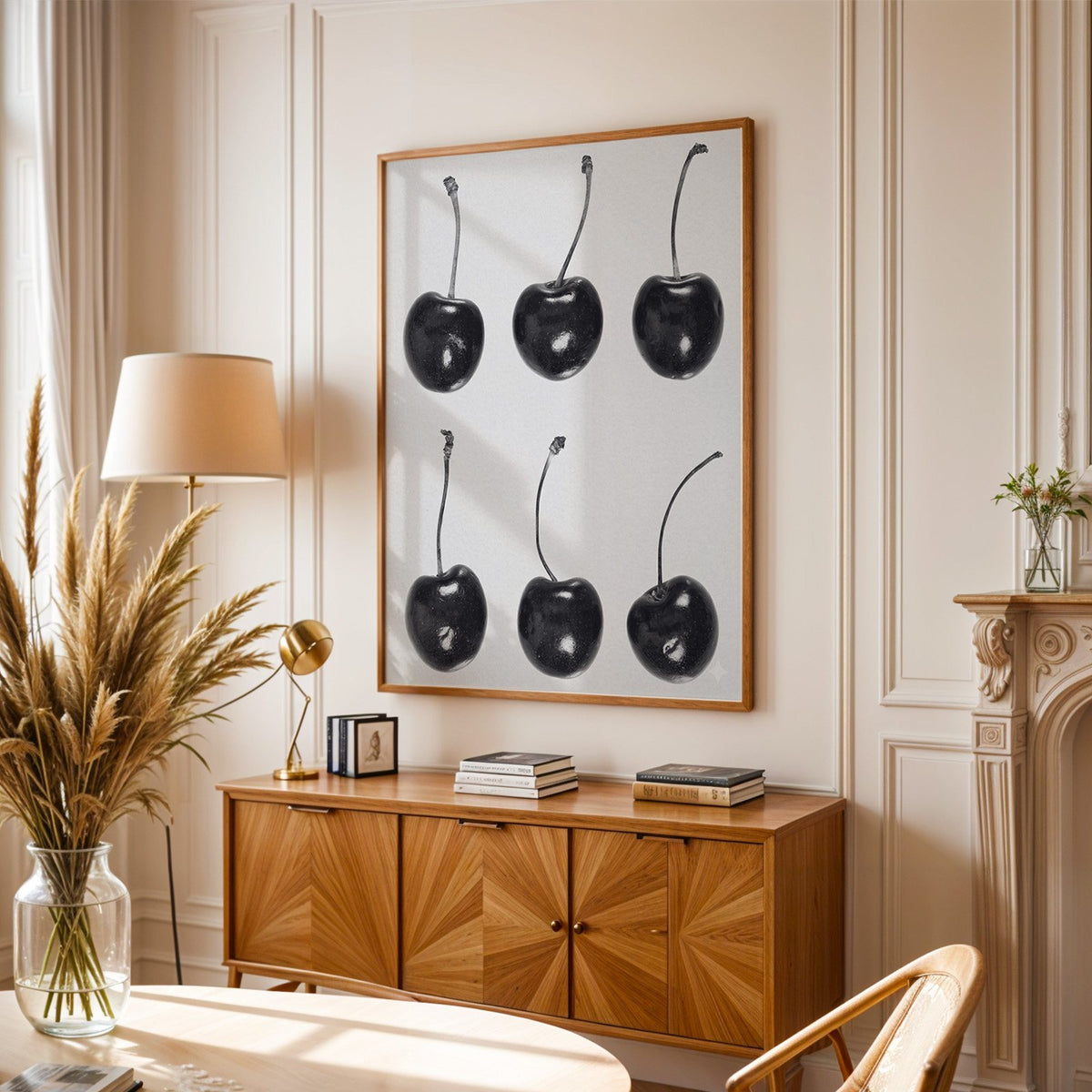 Six glossy black cherries in a grid on gray print in an oak frame above a sunburst wood sideboard with pampas grass