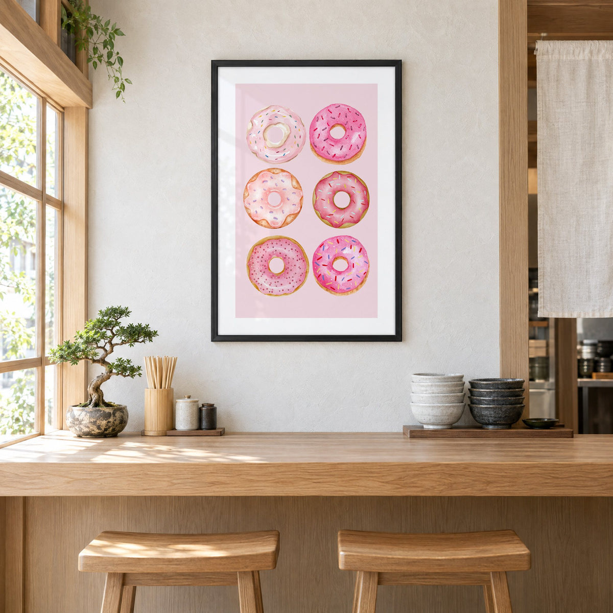 Six pink watercolor donuts print in a black frame above a light wood ramen counter with stools and stacked bowls