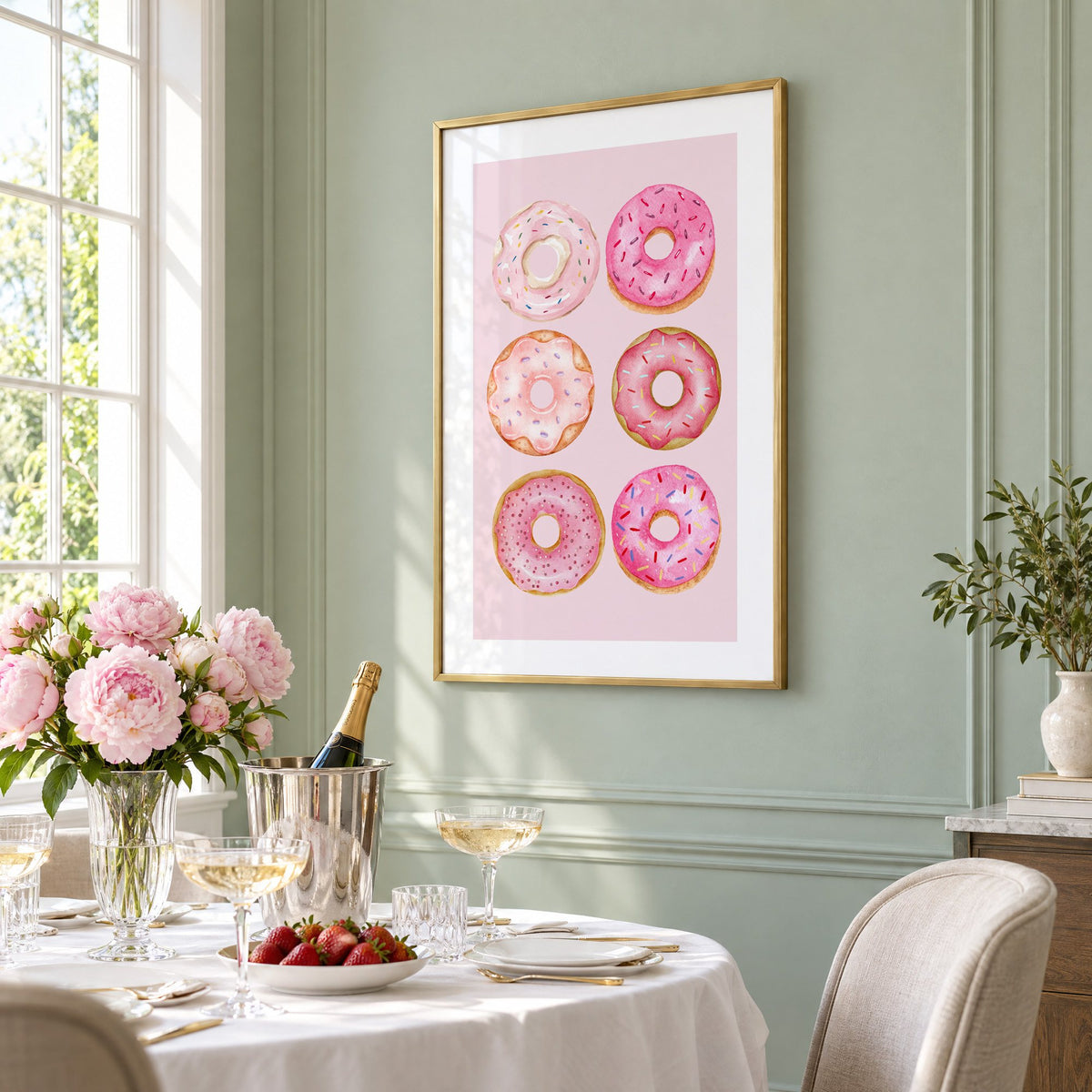 Six pink watercolor donuts print in a brass frame on a sage wall above a brunch table with champagne and peonies, seen at an angle