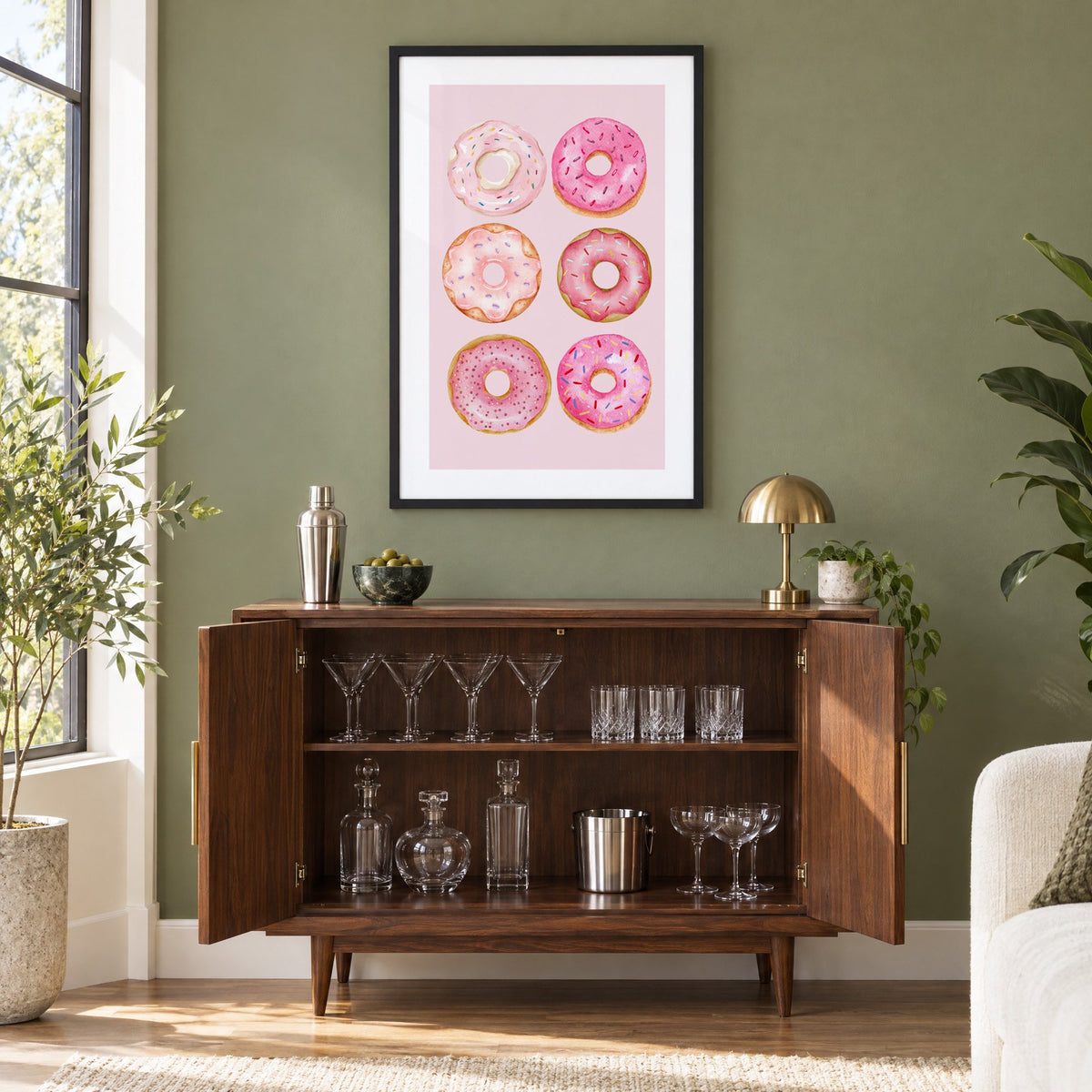 Six pink watercolor donuts print in a black frame on an olive green wall above a walnut bar cabinet with glassware