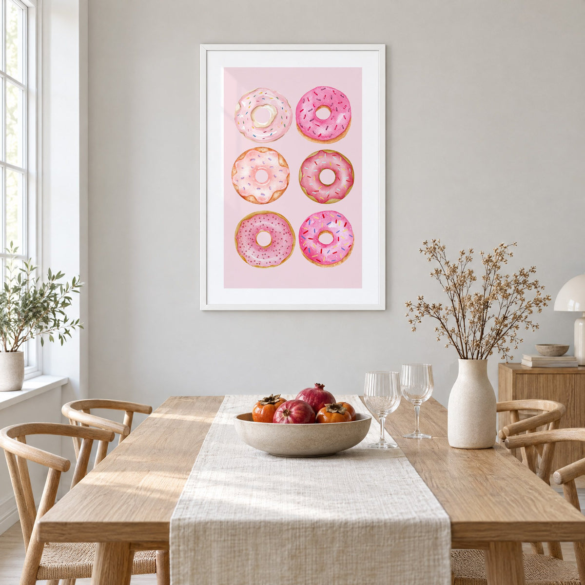 Six pink watercolor donuts print in a white frame above a light oak dining table with a linen runner and fruit bowl