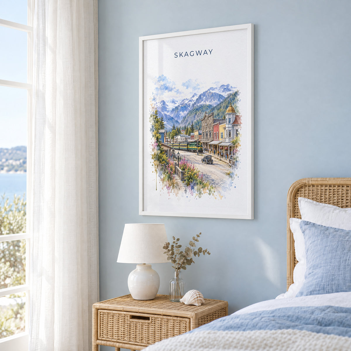 Skagway, Alaska watercolor print in a white frame on a pale blue wall beside a rattan bed in a coastal bedroom