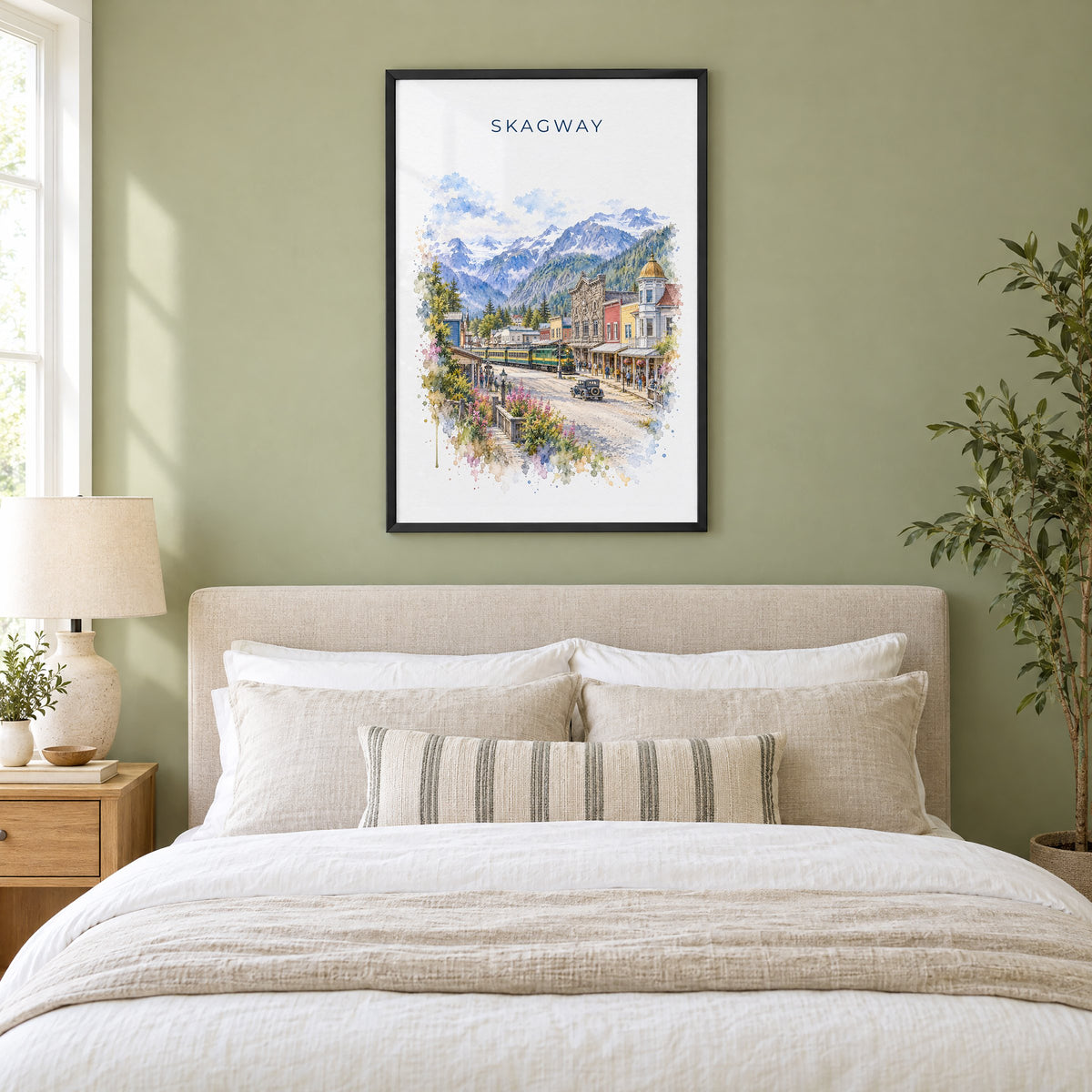 Skagway, Alaska watercolor print in a black frame above an upholstered headboard in a sage green bedroom