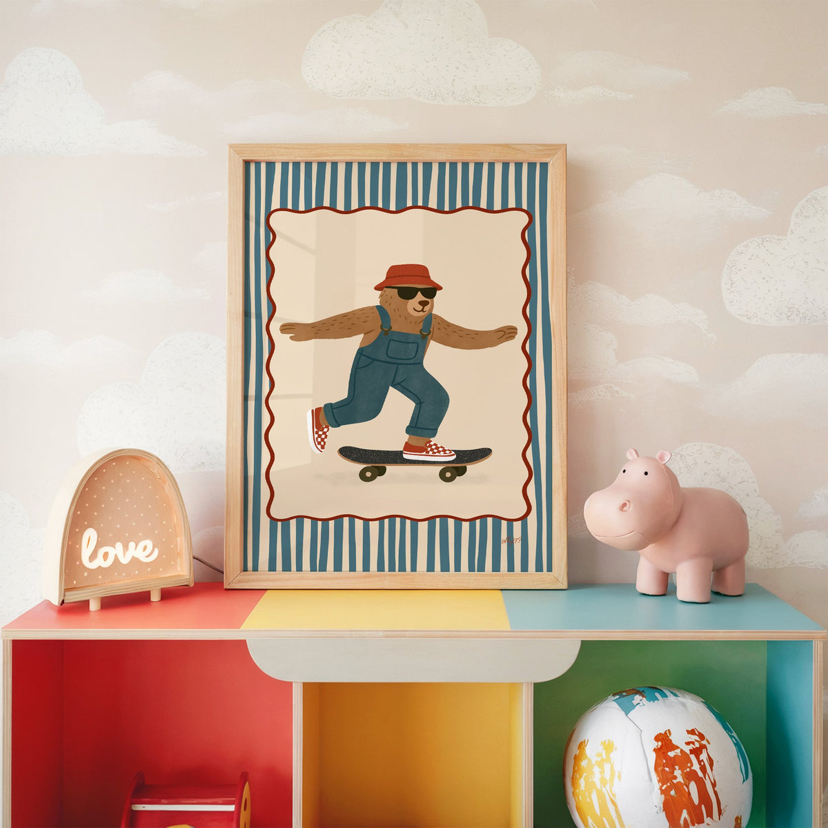 Bear in overalls on a skateboard nursery print in an oak frame on a rainbow toy shelf against cloud wallpaper