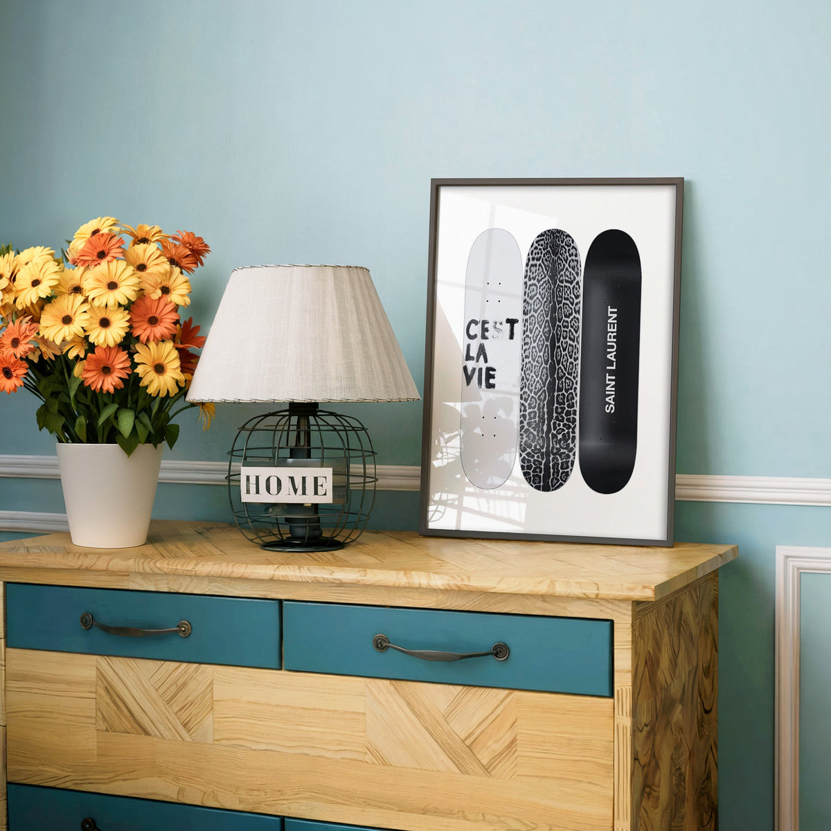 Three skateboards with leopard deck and lettering print in a charcoal frame on a teal dresser beside orange daisies