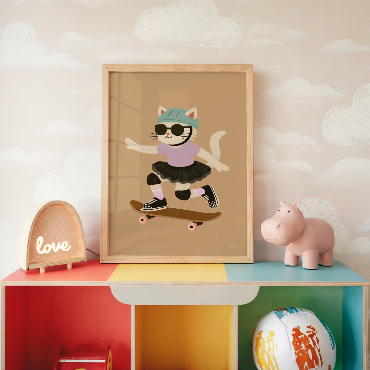 Skater cat in black tutu nursery print in an oak frame on a rainbow toy shelf against cloud wallpaper