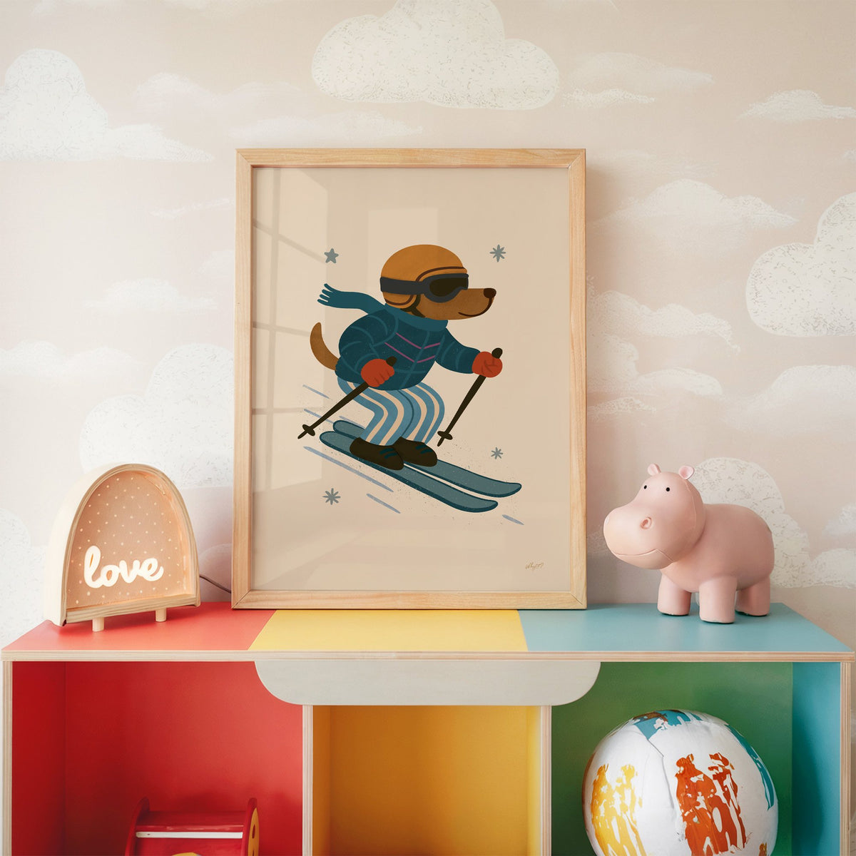 Skiing dog in puffer jacket nursery print in an oak frame on a rainbow toy shelf against cloud wallpaper