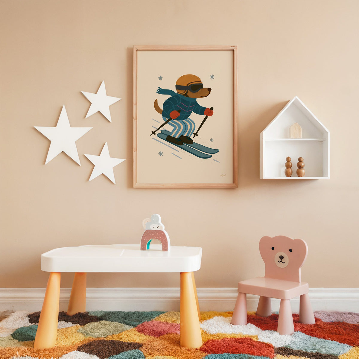Skiing dog in puffer jacket nursery print in an oak frame on a peach wall above a kids table with wall stars