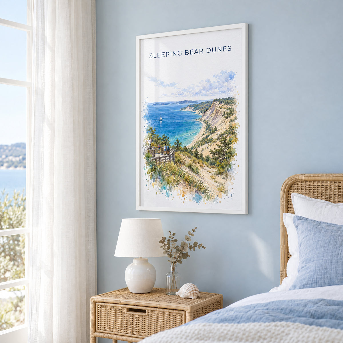 Sleeping Bear Dunes watercolor print in a white frame on a pale blue wall beside a rattan bed in a coastal bedroom