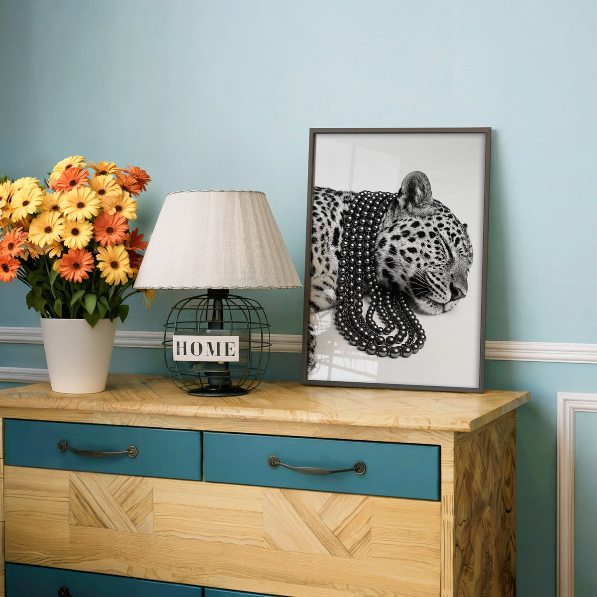 Sleeping leopard wearing black pearl strands print in a charcoal frame on a teal dresser beside orange daisies