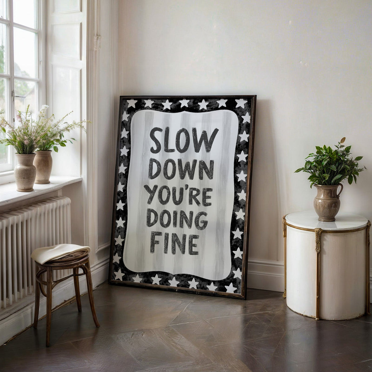 Slow down you're doing fine quote with star border print in a black frame on a dark stone floor beside a white side table