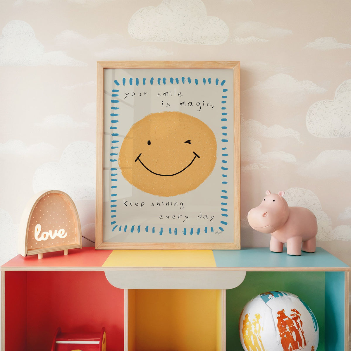 Winking smiley face quote nursery print in an oak frame on a rainbow toy shelf against cloud wallpaper