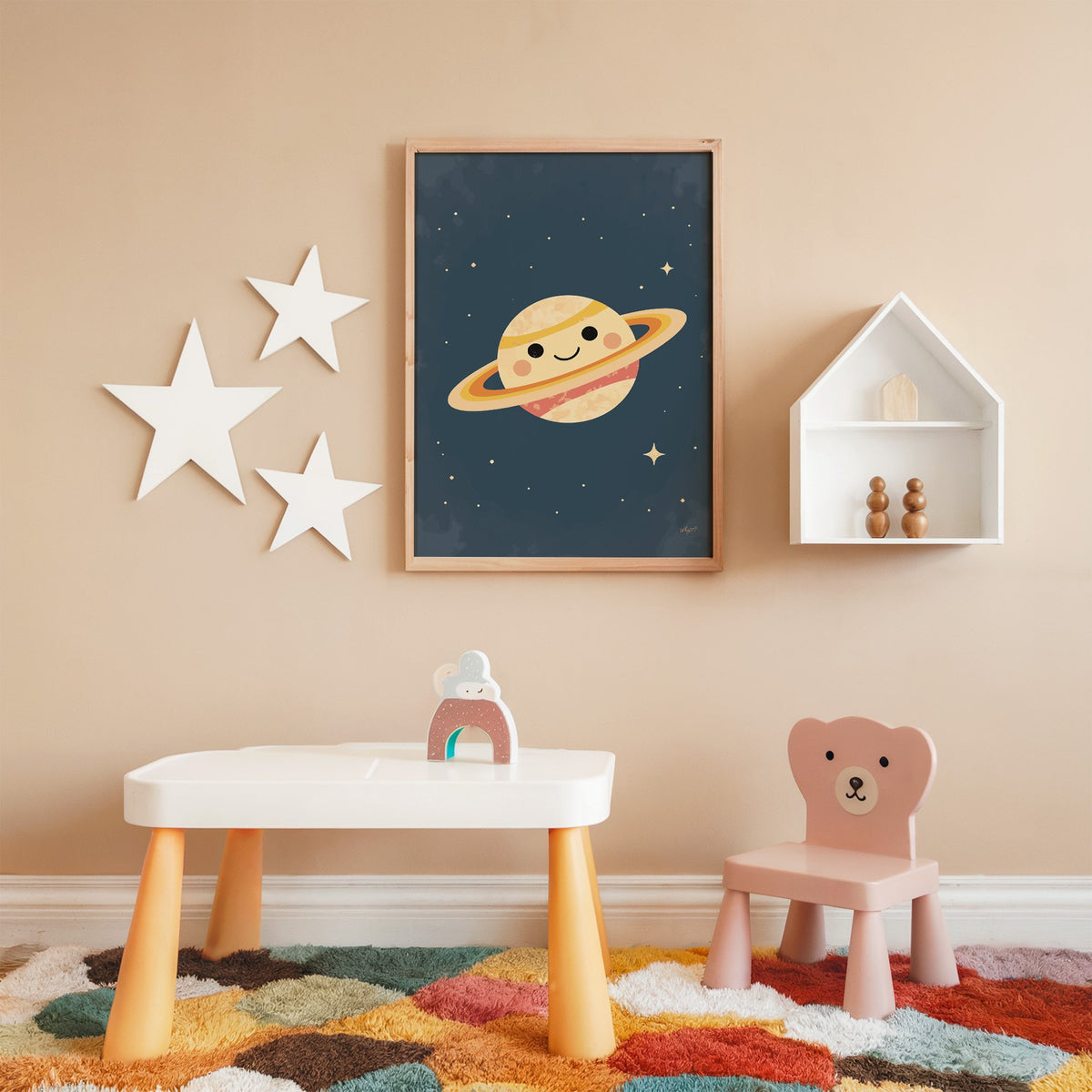 Smiling saturn with rosy cheeks nursery print in an oak frame on a peach wall above a kids table with wall stars