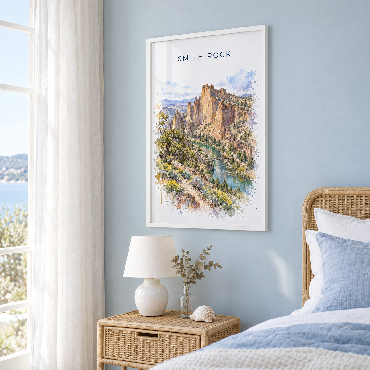 Smith Rock watercolor print in a white frame on a pale blue wall beside a rattan bed in a coastal bedroom
