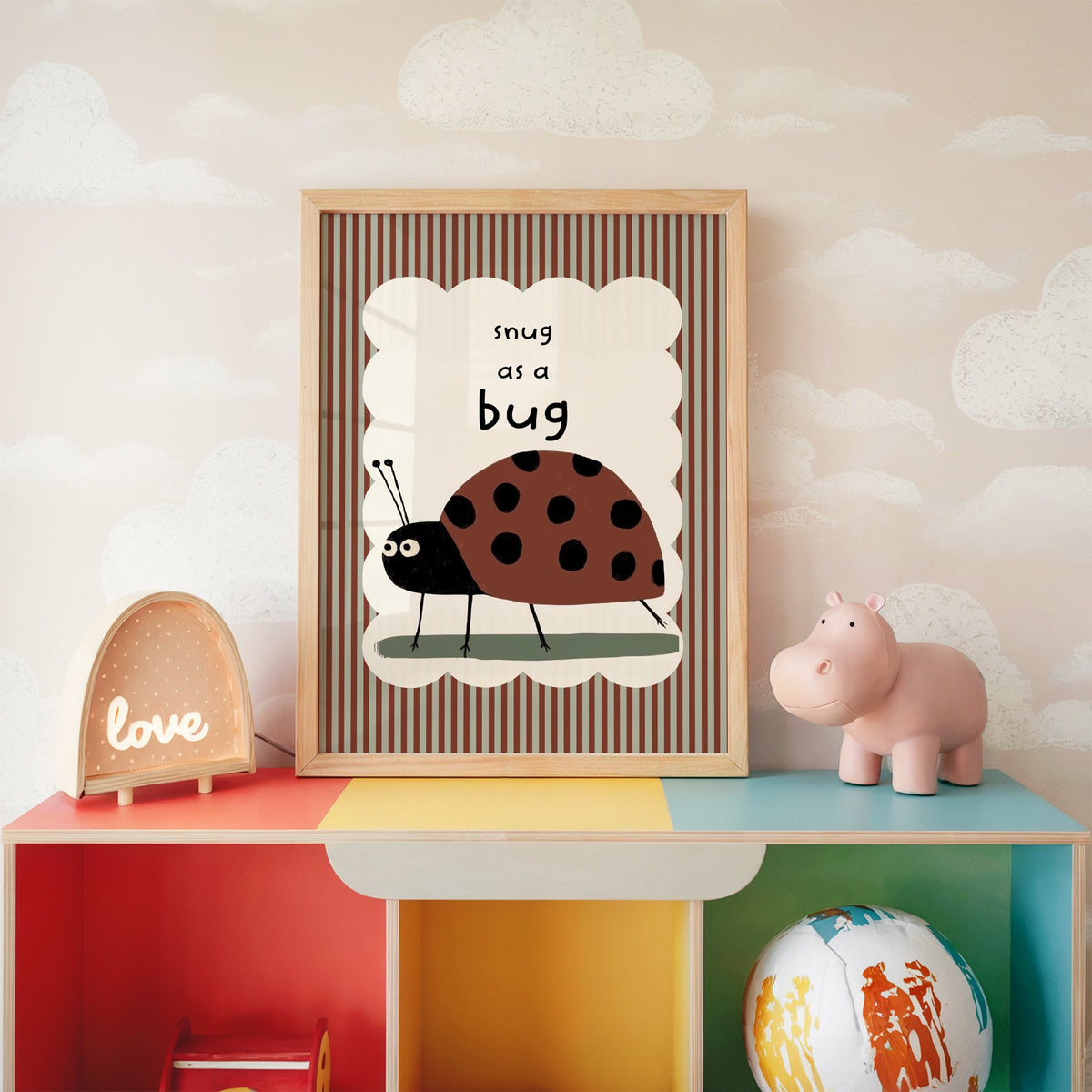 Snug as a bug ladybug nursery print in an oak frame on a rainbow toy shelf against cloud wallpaper