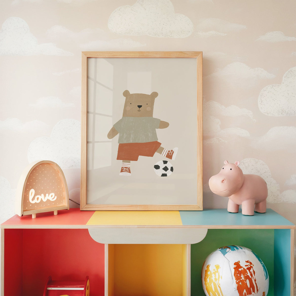Soccer bear orange shorts nursery print in an oak frame on a rainbow toy shelf against cloud wallpaper