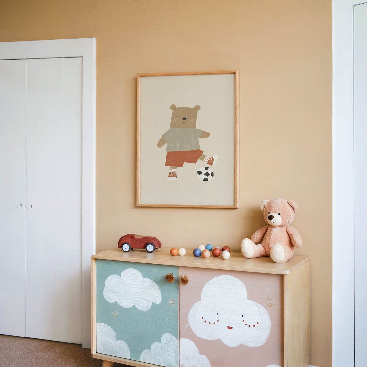 Soccer bear orange shorts nursery print in an oak frame on a peach wall above a cloud-painted cabinet