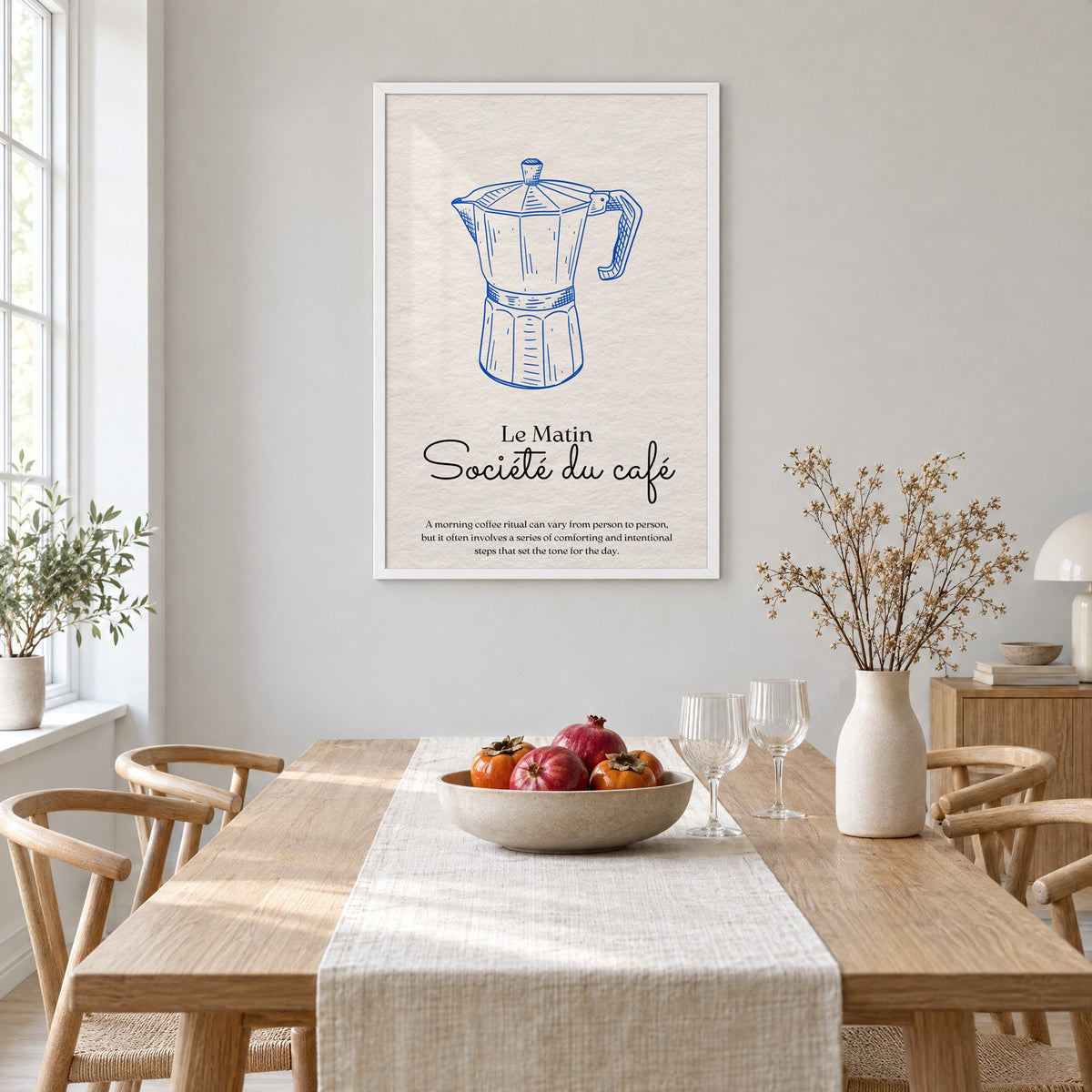Societe du cafe cobalt moka pot print in a white frame above a light oak dining table with a linen runner and fruit bowl