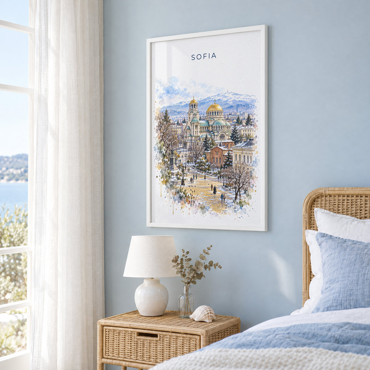 Sofia, Bulgaria watercolor print in a white frame on a pale blue wall beside a rattan bed in a coastal bedroom