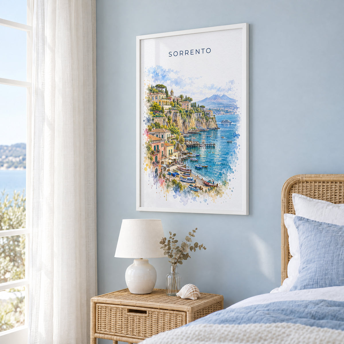 Sorrento watercolor print in a white frame on a pale blue wall beside a rattan bed in a coastal bedroom