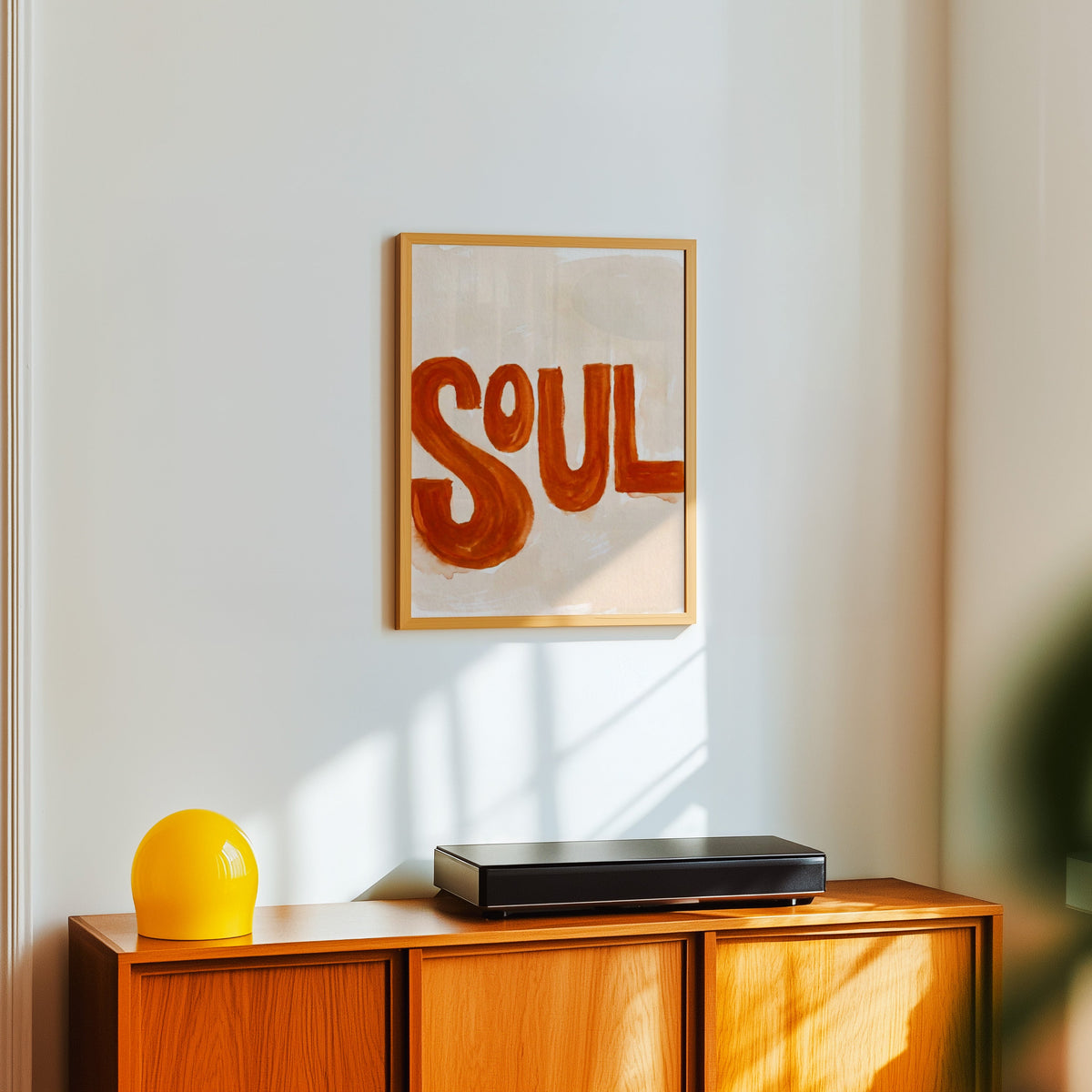 Soul brush lettering print in a light oak frame above a teak sideboard with a yellow lamp