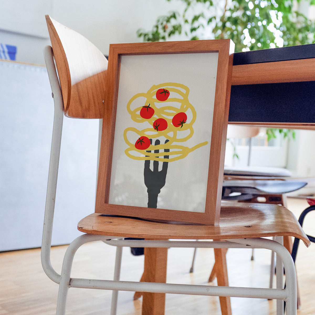 Spaghetti fork and cherry tomatoes print in a walnut frame resting on a plywood studio chair beside a dark desk