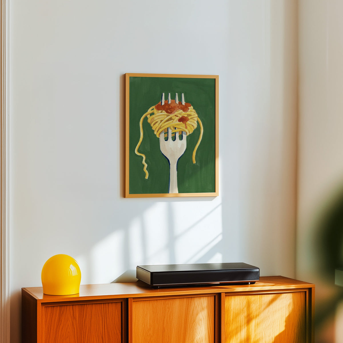 Green pasta wall art print in a light oak frame above a teak sideboard with a yellow lamp