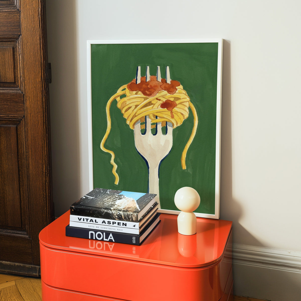 Watercolor pasta print in a white frame on a glossy orange cabinet with books
