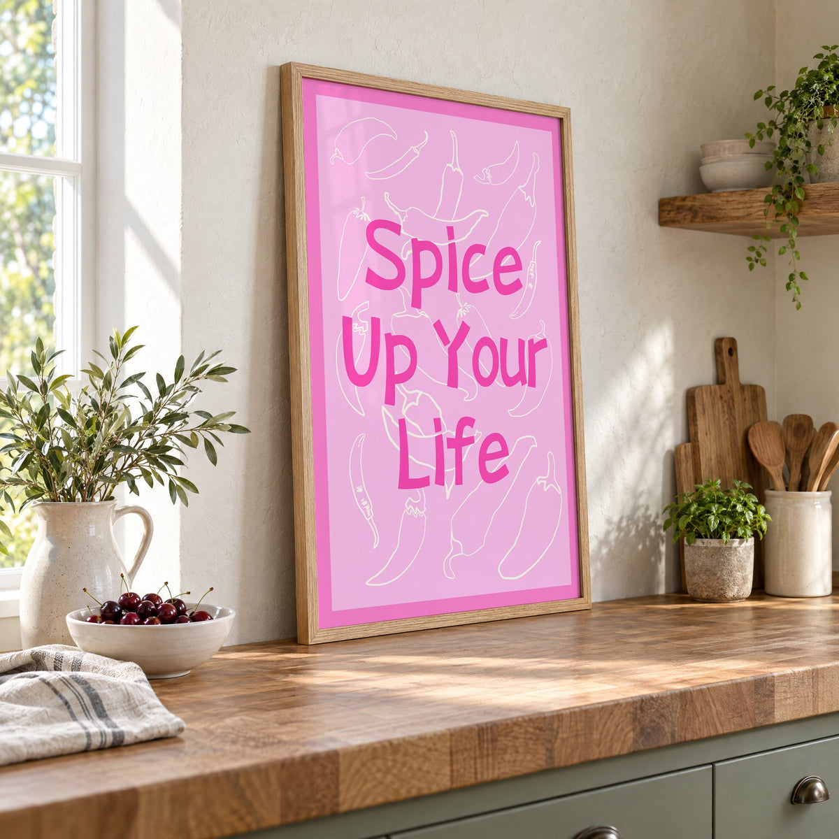 Spice Up Your Life pink chilli print in an oak frame leaning on a butcher block island beside a bowl of cherries