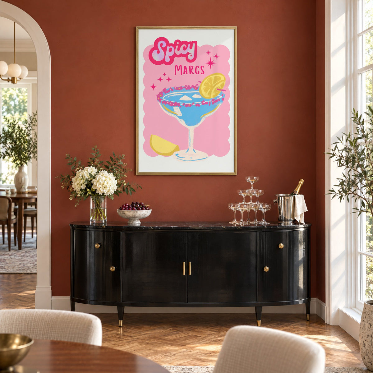 Spicy Margs blue margarita print in a brass frame on a terracotta wall above a black buffet with a champagne tower