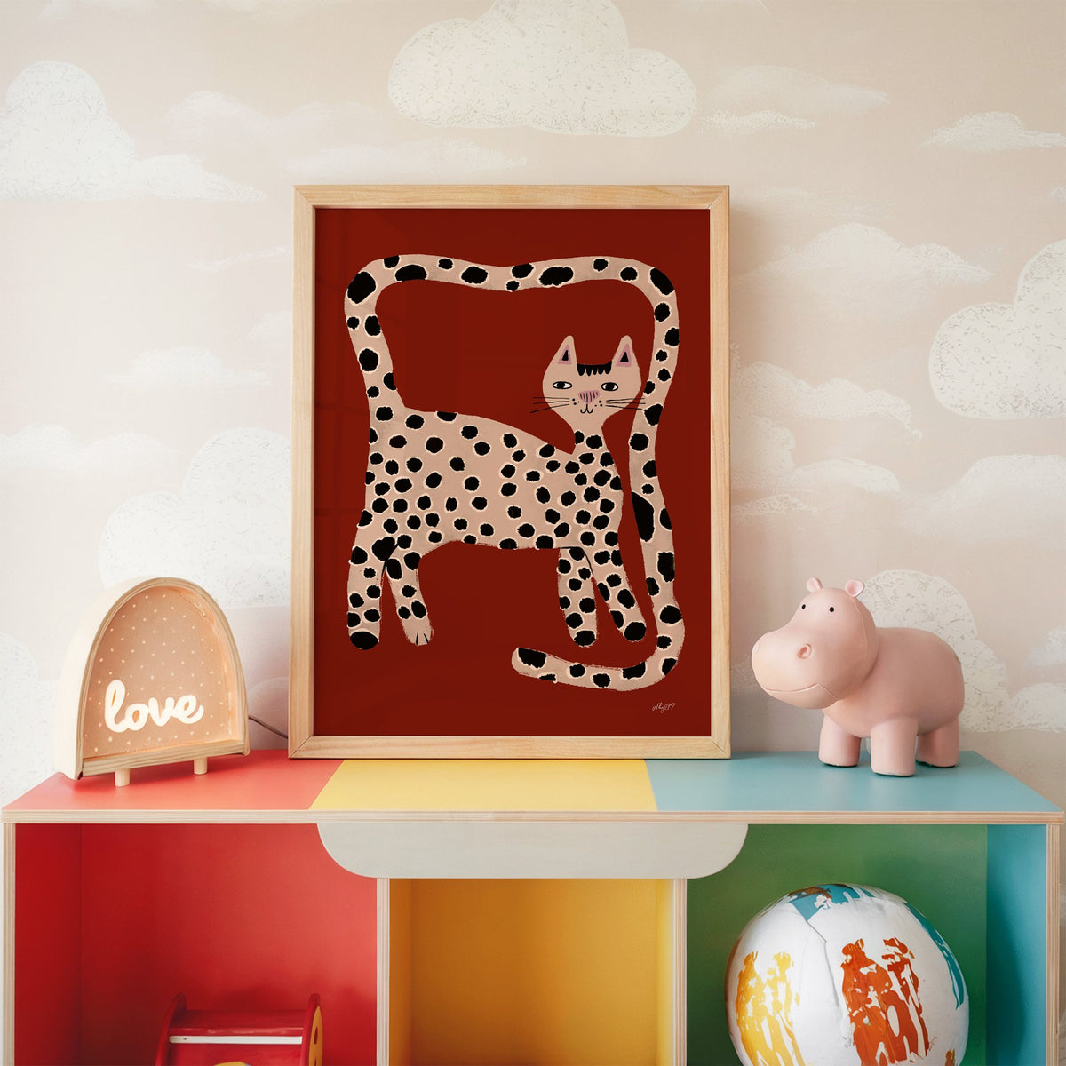 Spotted cat with looping tail nursery print in an oak frame on a rainbow toy shelf against cloud wallpaper