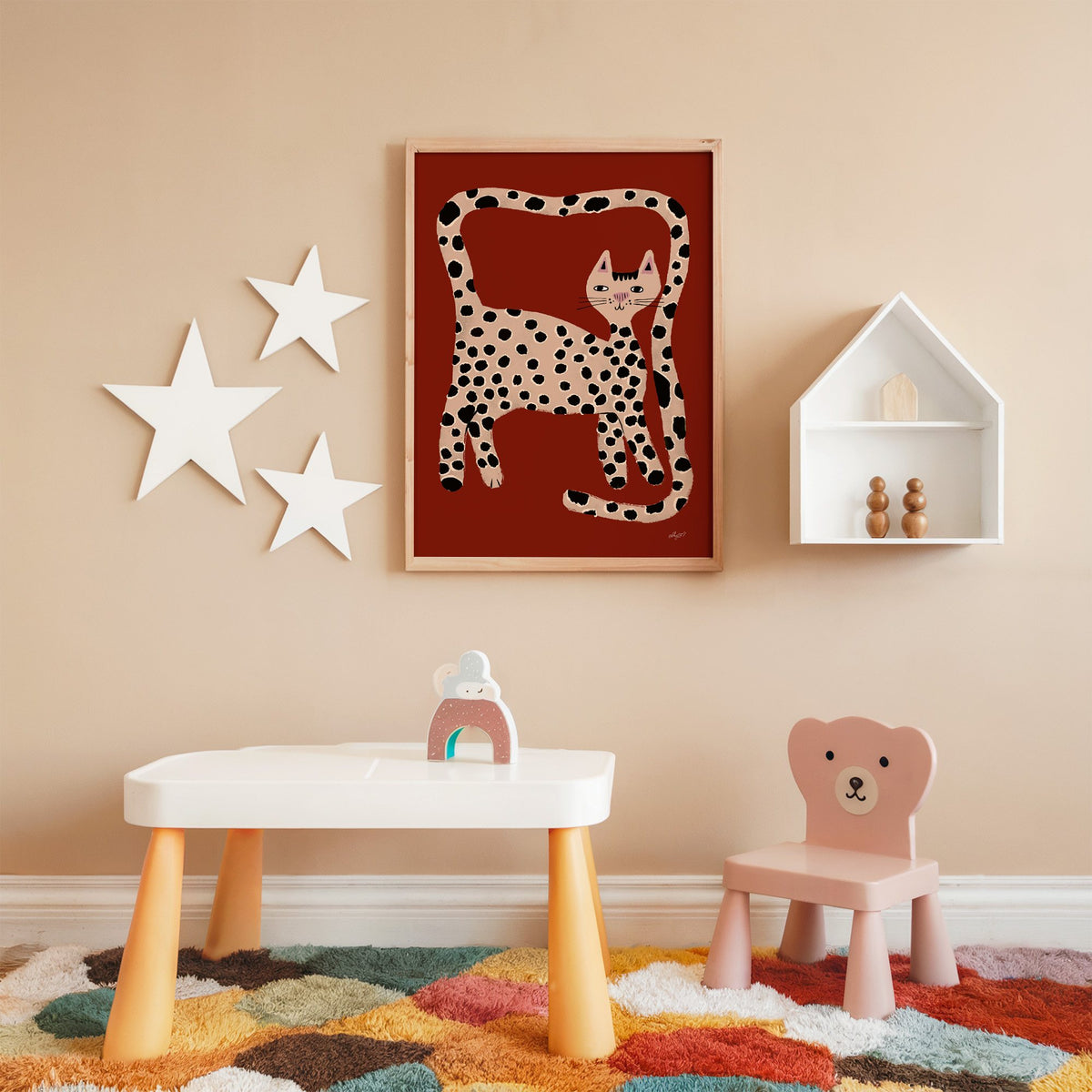 Spotted cat with looping tail nursery print in an oak frame on a peach wall above a kids table with wall stars