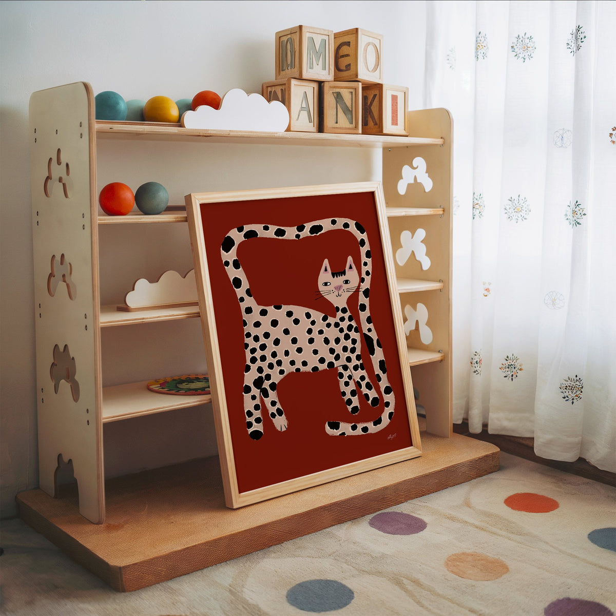 Spotted cat with looping tail nursery print in an oak frame leaning on a toy shelf with wooden letter blocks