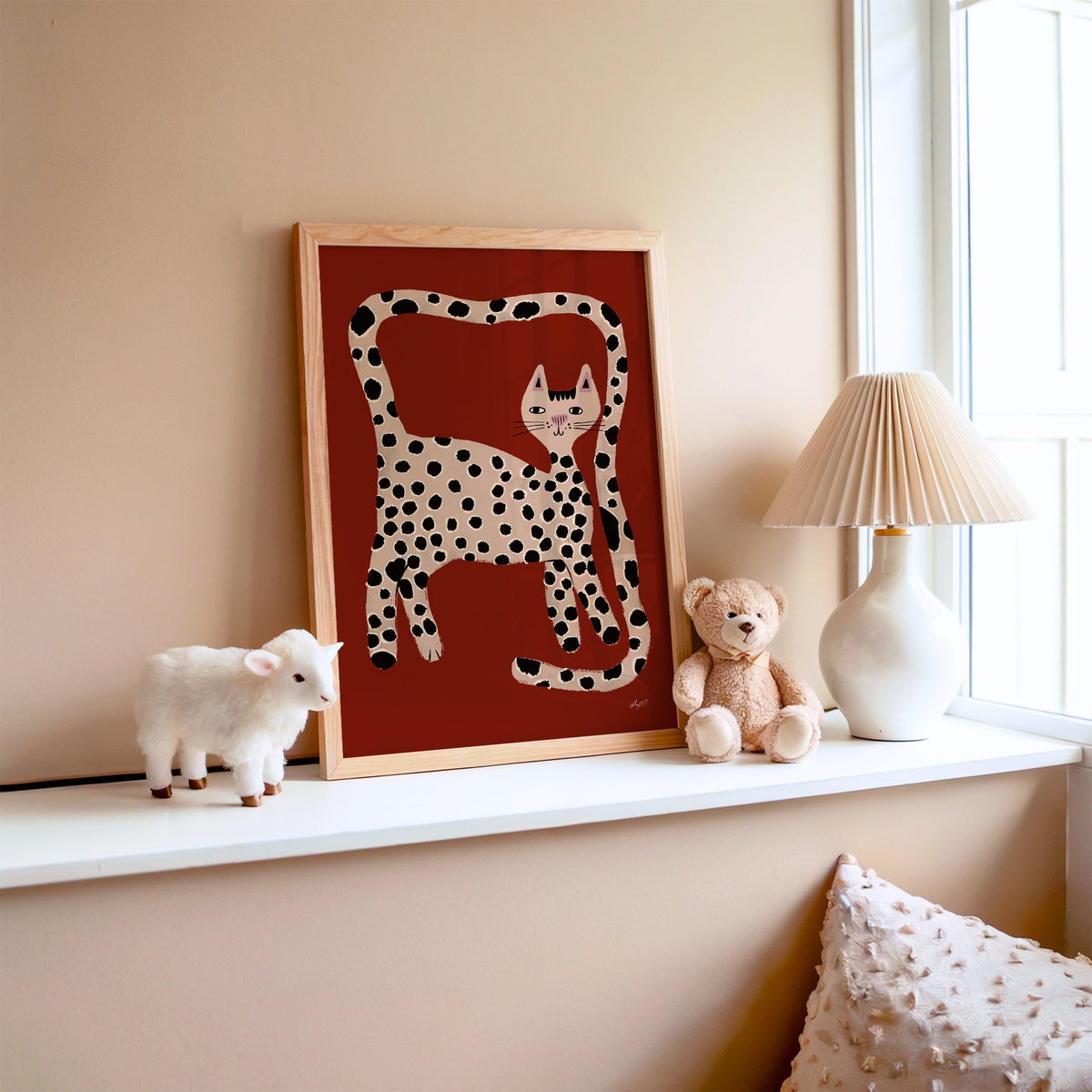 Spotted cat with looping tail nursery print in an oak frame on a window ledge with a toy lamb and teddy