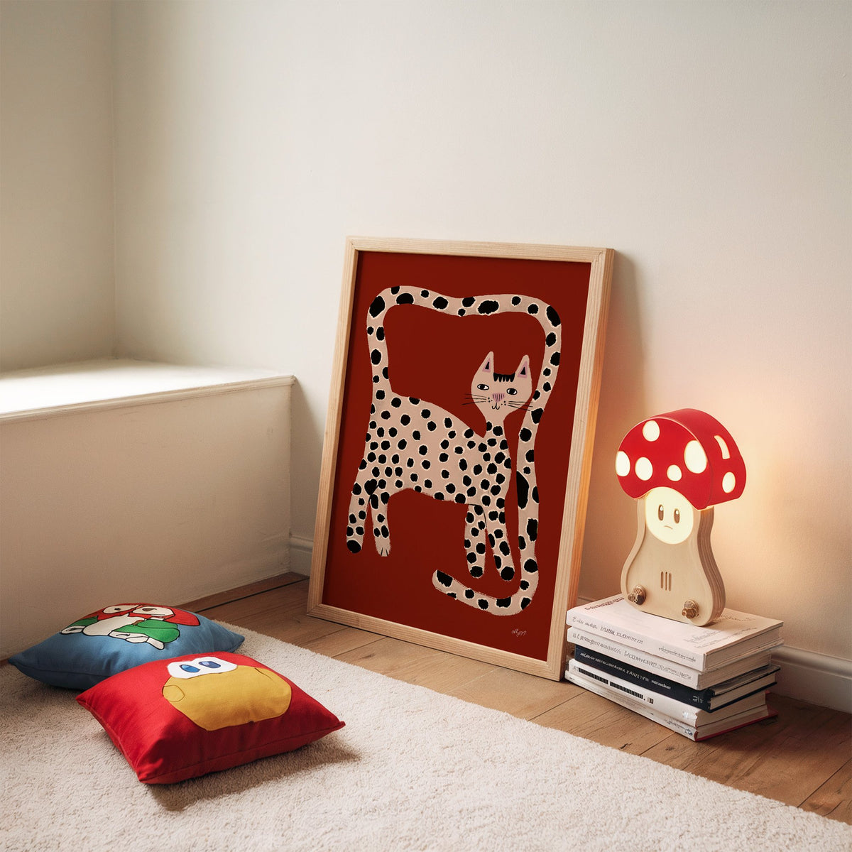 Spotted cat with looping tail nursery print in an oak frame leaning on a rug beside a mushroom lamp and floor cushions
