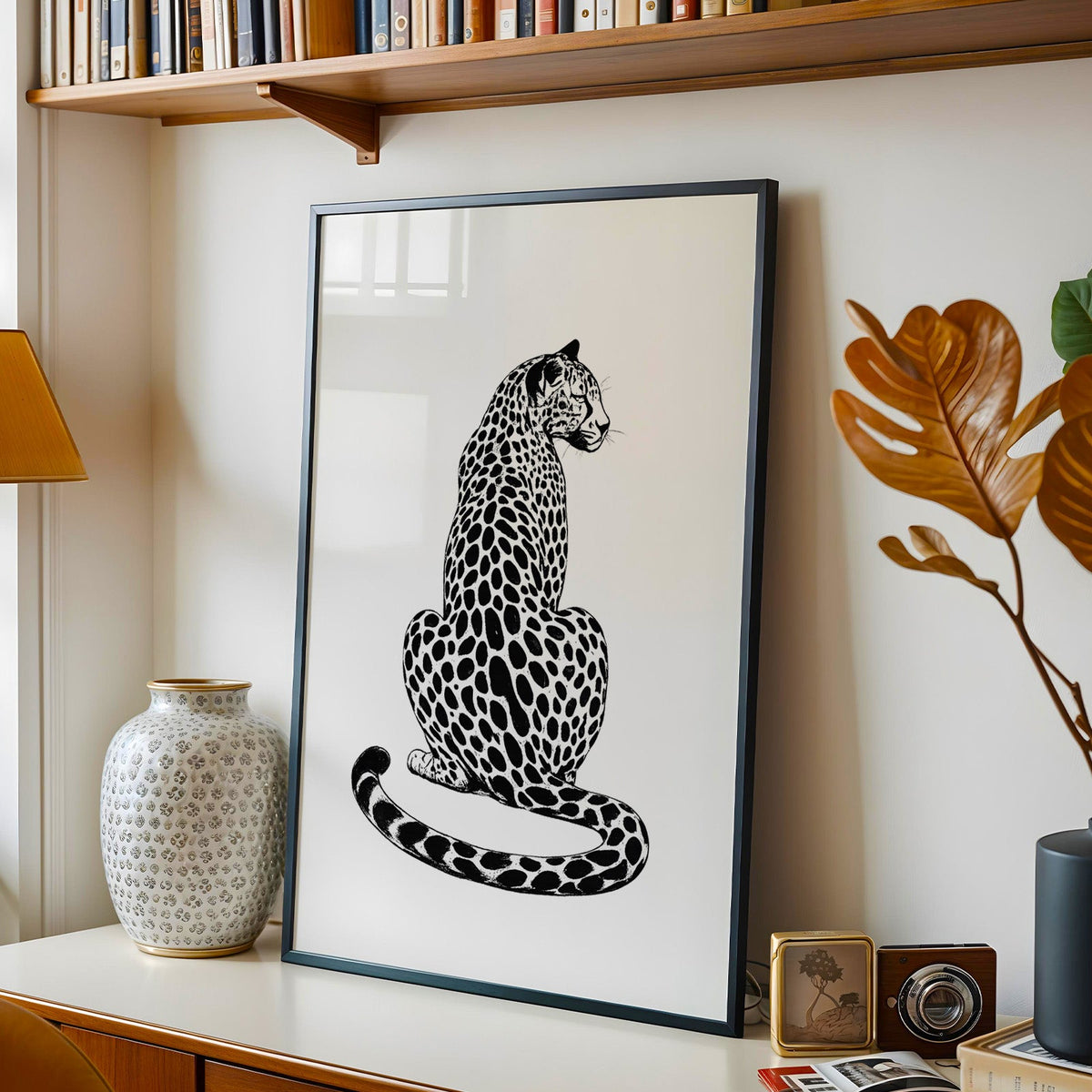 Spotted big cat seen from behind in black ink print in a black frame on a console under a bookshelf