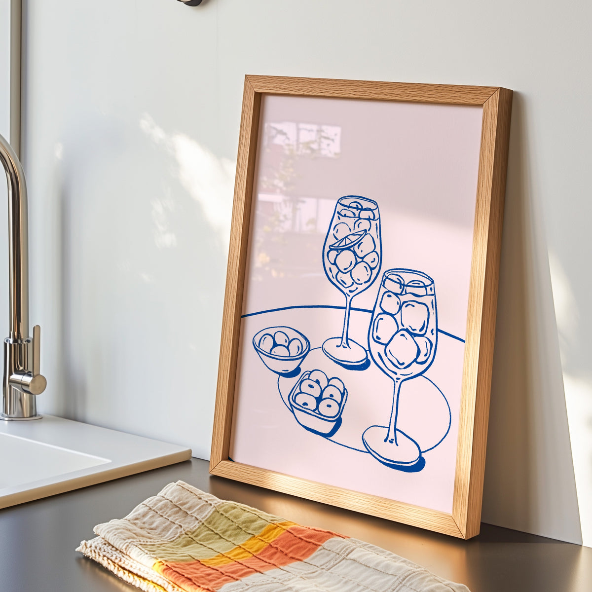 Spritz and olives print in an oak frame by the sink with a striped tea towel